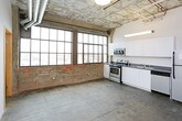 This single-floor urban loft masterfully blends industrial heritage with modern living. The open layout is defined by striking polished concrete floors that run throughout, exposed concrete ceilings with visible ductwork and conduit, and original brick accent walls that speak to the building's storied past. Oversized, multi-pane industrial windows flood the living and kitchen areas with natural light while offering expansive views of the surrounding cityscape.

The kitchen is well-appointed with white cabinetry, dark countertops, and a full suite of stainless steel appliances including a range, microwave, dishwasher, and refrigerator. The open kitchen flows into a generous living area, making the space ideal for both daily living and entertaining.

The sleeping quarters feature sliding wood-panel closet doors and clean white walls that provide a calm contrast to the raw industrial elements found elsewhere. Exposed brick columns and high ceilings carry through, maintaining the loft's architectural character.

Two bathrooms serve the space, each featuring a walk-in shower with white surrounds, a toilet, and polished fixtures. One bathroom includes a vanity with a vessel sink and mirror. Throughout the unit, the interplay of raw materials and modern finishes creates a dynamic, warehouse-inspired ambiance.