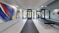 Located at 78 Braemar Dr in Brampton, Ontario, this building lobby serves as a polished and well-appointed entry point for residents and visitors alike. The space is designed with a clean, modern aesthetic, featuring large-format ceramic tile flooring in a neutral tone that runs the length of the corridor, complemented by a grey carpet runner leading from the glass entry doors inward. The entry vestibule is framed by floor-to-ceiling glass panels and a set of automatic black-framed glass doors, allowing natural light to filter into the interior and offering views of the surrounding outdoor greenery. Security cameras are positioned at the entrance, reflecting the building's attention to safety. A standout feature of the lobby is the prominent display of two large, vibrant abstract paintings mounted on the textured grey wall — their bold, colourful compositions adding a striking artistic focal point to the space. On the opposite side, a full-length mirror enhances the sense of depth and openness. A sleek black leather bench provides a functional seating area for waiting guests. Toward the rear of the lobby, stainless steel elevator doors are set against a refined stone-tile accent wall, providing access to the building's upper floors. Recessed ceiling lighting throughout ensures the space is evenly and warmly illuminated.