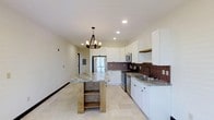 This single-floor space is highlighted by a thoughtfully designed kitchen that serves as its central feature. The kitchen showcases crisp white shaker-style cabinetry that extends to the ceiling, providing generous storage throughout. A striking freestanding island with a natural wood base and granite countertop anchors the space, complemented by an elegant multi-bulb chandelier suspended above it. Rich, dark tile backsplash creates a bold contrast against the white cabinetry, adding depth and visual interest to the room. Stainless steel appliances, including a refrigerator, range, and built-in microwave, lend a clean, contemporary finish. The undermount sink is paired with a matte black faucet, offering a refined touch. Light-toned large-format tile flooring runs throughout, enhancing the sense of openness and reflecting the ample recessed lighting overhead. The layout is open and functional, allowing for easy movement and a seamless flow between spaces. The overall ambiance is bright and inviting, combining modern finishes with warm, rustic accents to create a balanced and welcoming environment.