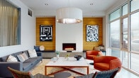 This thoughtfully designed single-floor lounge space makes a striking first impression through its cohesive mid-century modern aesthetic and carefully curated interiors. The focal point of the main seating area is a sleek linear fireplace set into a crisp white wall, flanked symmetrically by warm horizontal wood-panel cladding that extends across the room, adding texture and natural warmth. Bold black-and-white abstract artwork is displayed against the wood panels, complementing the vibrant orange lounge chairs, charcoal sofa, and woven accent seating arranged around a marble-topped coffee table. A large drum pendant light anchors the seating area from above, casting a warm glow over the space. Built-in bench seating with cushions runs along the wood-paneled wall, offering additional casual seating with understated style. Polished concrete flooring flows through the entry and corridor areas, transitioning seamlessly into tiled surfaces in the lounge. Floor-to-ceiling glazed windows along one side of the lounge flood the space with abundant natural light and provide views to the exterior. A glass-partitioned area with sheer curtaining offers a defined, semi-private zone within the open layout. The entry corridor features elevator access and curated framed artwork, reinforcing the space's gallery-like attention to design detail throughout.