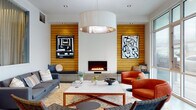 This thoughtfully designed single-floor lounge space makes a striking first impression through its cohesive mid-century modern aesthetic and carefully curated interiors. The focal point of the main seating area is a sleek linear fireplace set into a crisp white wall, flanked symmetrically by warm horizontal wood-panel cladding that extends across the room, adding texture and natural warmth. Bold black-and-white abstract artwork is displayed against the wood panels, complementing the vibrant orange lounge chairs, charcoal sofa, and woven accent seating arranged around a marble-topped coffee table. A large drum pendant light anchors the seating area from above, casting a warm glow over the space. Built-in bench seating with cushions runs along the wood-paneled wall, offering additional casual seating with understated style. Polished concrete flooring flows through the entry and corridor areas, transitioning seamlessly into tiled surfaces in the lounge. Floor-to-ceiling glazed windows along one side of the lounge flood the space with abundant natural light and provide views to the exterior. A glass-partitioned area with sheer curtaining offers a defined, semi-private zone within the open layout. The entry corridor features elevator access and curated framed artwork, reinforcing the space's gallery-like attention to design detail throughout.