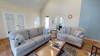 This single-floor space offers a comfortable and well-appointed interior with a thoughtfully designed layout. The living area is the centerpiece of the space, characterized by dramatic vaulted ceilings that create an impressive sense of height and openness. Warm natural light pours in through multiple windows and a glass-paneled door, illuminating the light-toned walls and hardwood flooring throughout.

The living area is furnished with two generous grey sofas accented with blue and white decorative pillows, arranged around a wooden coffee table with a lower storage shelf — a practical and stylish focal point for the room. A patterned area rug anchors the seating arrangement, adding texture and warmth to the space.

Beyond the living area, a glass-paned interior door leads to an adjacent room, offering a sense of connected but distinct spaces within the floor plan. A wall-mounted clock and a recessed niche add subtle architectural character to the walls, while the overall color palette — soft whites, warm neutrals, and natural wood tones — contributes to a calm and inviting atmosphere. The abundance of natural light and the airy ceiling height make this a bright, pleasant space suitable for relaxed everyday living.