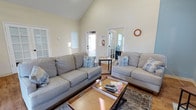 This single-floor space offers a comfortable and well-appointed interior with a thoughtfully designed layout. The living area is the centerpiece of the space, characterized by dramatic vaulted ceilings that create an impressive sense of height and openness. Warm natural light pours in through multiple windows and a glass-paneled door, illuminating the light-toned walls and hardwood flooring throughout.

The living area is furnished with two generous grey sofas accented with blue and white decorative pillows, arranged around a wooden coffee table with a lower storage shelf — a practical and stylish focal point for the room. A patterned area rug anchors the seating arrangement, adding texture and warmth to the space.

Beyond the living area, a glass-paned interior door leads to an adjacent room, offering a sense of connected but distinct spaces within the floor plan. A wall-mounted clock and a recessed niche add subtle architectural character to the walls, while the overall color palette — soft whites, warm neutrals, and natural wood tones — contributes to a calm and inviting atmosphere. The abundance of natural light and the airy ceiling height make this a bright, pleasant space suitable for relaxed everyday living.