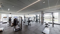 Located at 315 First Ave NE in Minneapolis, MN, this well-appointed fitness center offers a spacious, single-floor layout designed to accommodate a full range of workout activities. The facility features a thoughtfully organized arrangement of professional-grade equipment, including strength-training machines, cable systems, free weights, ellipticals, stationary bikes, and treadmills, providing users with a comprehensive exercise experience.

The interior design is contemporary and polished, with a paneled ceiling accented by linear LED lighting strips that cast a clean, even illumination throughout the space. The durable rubber flooring provides both safety and functionality, ideal for high-traffic fitness use. Structural concrete columns are integrated seamlessly into the design, adding an industrial aesthetic that complements the modern finishes.

Floor-to-ceiling windows line multiple walls, flooding the space with natural light and offering open views of the surrounding Minneapolis landscape, creating an energizing atmosphere for workouts. A large-format display screen is mounted on one wall, adding a multimedia element to the environment. The overall ambiance is bright, clean, and professional, making this fitness center a well-equipped destination for residents or users seeking a complete training facility.