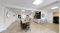 This single-floor community space is thoughtfully designed to serve as a versatile gathering hub. The main hall is expansive and open, featuring light wood-style flooring, recessed skylight panels, and a textured ceiling that together create a bright, welcoming atmosphere. A long communal dining table with industrial-style metal chairs anchors the central area, making it ideal for large group gatherings or events. Decorative bird paintings adorn the white walls, adding an artistic touch throughout.

Adjacent to the main hall, a secondary dining area offers multiple round tables with upholstered chairs, natural light from slatted windows, and structural columns that define the space without closing it off. A built-in wet bar station features dark subway tile backsplash, open shelving, and a sink — perfect for hosting and catering needs.

The lounge area provides a bold contrast with deep navy-blue walls, vivid artwork, yellow accent chairs, and a charcoal sofa arranged over a woven jute rug. Statement globe sconces and a lush fiddle-leaf fig plant complete the inviting, curated aesthetic of this room.

A dedicated on-site laundry facility houses multiple stacked washer and dryer units, finished with durable tile flooring for easy maintenance. Together, these spaces form a well-appointed, amenity-rich community center.