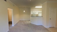 This single-floor unit features an open-concept design that connects the living area and kitchen seamlessly. The living space is finished with neutral-toned wall-to-wall carpeting and painted in warm beige tones, creating a clean and understated atmosphere. Crown molding runs along the upper edges of the walls, adding a subtle architectural detail throughout. A pendant light fixture hangs centrally, providing focused illumination over the main living area.

The kitchen is positioned toward the rear and defined by a half-wall peninsula with a dark countertop, offering both a natural division between spaces and a casual surface for dining or meal preparation. White cabinetry lines the upper walls of the kitchen, providing ample storage, while a white refrigerator, microwave, and range complete the appliance suite. A fluorescent light fixture above the kitchen ensures well-lit functionality during food preparation.

The overall layout is practical and efficient, with consistent finishes throughout that contribute to a cohesive aesthetic. The neutral palette and open flow between the kitchen and living area make the space feel airy and adaptable for various furnishing arrangements.