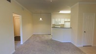 This single-floor unit features an open-concept design that connects the living area and kitchen seamlessly. The living space is finished with neutral-toned wall-to-wall carpeting and painted in warm beige tones, creating a clean and understated atmosphere. Crown molding runs along the upper edges of the walls, adding a subtle architectural detail throughout. A pendant light fixture hangs centrally, providing focused illumination over the main living area.

The kitchen is positioned toward the rear and defined by a half-wall peninsula with a dark countertop, offering both a natural division between spaces and a casual surface for dining or meal preparation. White cabinetry lines the upper walls of the kitchen, providing ample storage, while a white refrigerator, microwave, and range complete the appliance suite. A fluorescent light fixture above the kitchen ensures well-lit functionality during food preparation.

The overall layout is practical and efficient, with consistent finishes throughout that contribute to a cohesive aesthetic. The neutral palette and open flow between the kitchen and living area make the space feel airy and adaptable for various furnishing arrangements.