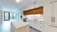 This contemporary single-floor residence showcases a thoughtfully designed interior defined by clean lines, a neutral palette, and high-quality finishes throughout. The standout feature is the chef's kitchen, which combines flat-front white cabinetry with warm wood upper cabinets, creating a striking two-tone contrast. A marble-look backsplash extends across the full wall, complementing the white quartz countertops. Stainless steel appliances, including a gas cooktop, wall oven, and built-in microwave, are seamlessly integrated into the cabinetry. A central island provides additional prep and storage space, while light-toned concrete flooring grounds the area. The open living space flows naturally from the kitchen and is bathed in natural light from floor-to-ceiling sliding glass doors that open to an outdoor area with urban views. Light hardwood flooring runs throughout the living and bedroom areas, adding warmth and continuity. The bedroom is generously sized and features direct access to an organized walk-in closet with built-in shelving. The bathroom is finished with large-format stone-look floor tiles, a herringbone-patterned feature wall, a floating wood vanity with an illuminated mirror, and a deep soaking tub — delivering a spa-like atmosphere that completes this refined, modern residence.