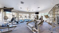 Located at 9971 E Speedway Blvd in Tucson, Arizona, this fitness center is a comprehensive, single-floor facility designed to accommodate a wide range of workout needs. The space is organized into distinct exercise zones, each thoughtfully appointed with modern equipment and functional finishes.

The cardio area is anchored by a row of treadmills and complemented by elliptical trainers and stationary bikes, all from True Fitness. Large windows line the walls, flooding the space with natural light and offering views of the surrounding property. A ceiling fan overhead and recessed lighting contribute to a comfortable workout environment.

The strength training zone features a variety of cable machines, multi-function weight stations, and bench press setups, providing ample options for resistance training. An adjacent free weights area includes a dumbbell rack, stability balls, and an adjustable workout bench, catering to functional and flexibility training as well.

Throughout the fitness center, wall-mounted flat-screen televisions are positioned for easy viewing from multiple workout stations. The flooring transitions between durable carpet tile and hardwood-style surfaces, reflecting a clean and practical design. Accent walls in soft green tones add a refreshing visual element to the otherwise neutral interior palette. The overall ambiance is bright, open, and purpose-built for an effective fitness experience.