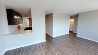 This single-floor space offers a clean, modern aesthetic defined by an open-concept layout that promotes a seamless flow between living areas. Dark hardwood-style flooring runs throughout, providing a rich, warm contrast against the crisp, neutral-toned walls and white baseboards. The spacious main living area feels airy and uncluttered, with a textured ceiling adding subtle character overhead. A half-wall partition with a pass-through counter connects the kitchen to the living space, creating an efficient yet social design. The kitchen is equipped with sleek cabinetry, a dark cooktop, and stainless steel appliances, including a microwave, all set against light countertops that brighten the space. A hallway visible toward the rear of the unit suggests additional rooms beyond, hinting at a thoughtfully organized floor plan. The overall ambiance is contemporary and understated, with a neutral color palette that offers a versatile canvas for a variety of interior styles. The space is well-lit with warm artificial lighting complementing the open layout throughout.