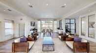 Located at 222 Kyser Blvd in Madison, Alabama, this single-story community clubhouse offers a thoughtfully designed gathering space that blends comfort, functionality, and contemporary style. The open-plan layout is anchored by a spacious lounge area featuring rich hardwood-style flooring, crown molding, and recessed lighting throughout. Structural columns add architectural interest while defining distinct zones within the open floor plan. The lounge is furnished with solid wood armchairs accented with cream upholstery and purple accent pillows, arranged around a sleek coffee table atop a neutral area rug. Large windows dressed with sheer curtains flood the interior with abundant natural light and offer views of the surrounding outdoor spaces. A dedicated business center includes a built-in dark countertop workstation with multiple computers, a wall-mounted display, and ample seating. Adjacent to the lounge, a fitness center is visible through striking black-framed glass partition walls, housing a variety of cardio and strength-training equipment. Private office spaces with glass-paneled doors provide quieter areas for meetings or focused work. A counter-height refreshment station completes the amenities. The overall ambiance is clean, bright, and welcoming, reflecting a modern residential community aesthetic.
