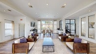 Located at 222 Kyser Blvd in Madison, Alabama, this single-story community clubhouse offers a thoughtfully designed gathering space that blends comfort, functionality, and contemporary style. The open-plan layout is anchored by a spacious lounge area featuring rich hardwood-style flooring, crown molding, and recessed lighting throughout. Structural columns add architectural interest while defining distinct zones within the open floor plan. The lounge is furnished with solid wood armchairs accented with cream upholstery and purple accent pillows, arranged around a sleek coffee table atop a neutral area rug. Large windows dressed with sheer curtains flood the interior with abundant natural light and offer views of the surrounding outdoor spaces. A dedicated business center includes a built-in dark countertop workstation with multiple computers, a wall-mounted display, and ample seating. Adjacent to the lounge, a fitness center is visible through striking black-framed glass partition walls, housing a variety of cardio and strength-training equipment. Private office spaces with glass-paneled doors provide quieter areas for meetings or focused work. A counter-height refreshment station completes the amenities. The overall ambiance is clean, bright, and welcoming, reflecting a modern residential community aesthetic.