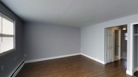 This single-floor space presents a straightforward, well-maintained interior with a clean and neutral aesthetic. The main room is finished with rich, dark hardwood flooring that contrasts attractively against the cool light-gray painted walls, creating a calm and versatile backdrop suitable for a variety of furnishing styles. White baseboards run along the perimeter of the room, adding a crisp, defined border between the walls and floor. The ceiling is smooth and painted white, contributing to a bright and open feel within the space. A doorway visible along one wall provides access to an adjacent area, suggesting a functional layout. The overall ambiance is minimalist and move-in ready, offering a blank canvas that allows future occupants to personalize the space to their preferences. The simplicity of the finishes ensures broad appeal, while the combination of hardwood flooring and neutral wall tones gives the space a timeless quality. The room benefits from an uncluttered design, making it easy to envision a range of interior arrangements and decor styles.