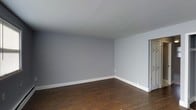 This single-floor space presents a straightforward, well-maintained interior with a clean and neutral aesthetic. The main room is finished with rich, dark hardwood flooring that contrasts attractively against the cool light-gray painted walls, creating a calm and versatile backdrop suitable for a variety of furnishing styles. White baseboards run along the perimeter of the room, adding a crisp, defined border between the walls and floor. The ceiling is smooth and painted white, contributing to a bright and open feel within the space. A doorway visible along one wall provides access to an adjacent area, suggesting a functional layout. The overall ambiance is minimalist and move-in ready, offering a blank canvas that allows future occupants to personalize the space to their preferences. The simplicity of the finishes ensures broad appeal, while the combination of hardwood flooring and neutral wall tones gives the space a timeless quality. The room benefits from an uncluttered design, making it easy to envision a range of interior arrangements and decor styles.