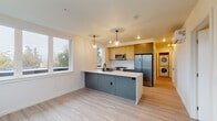 This single-floor unit showcases a clean, contemporary design with light wood-tone flooring running throughout all rooms, creating a cohesive and airy feel. The open-concept kitchen and living area serves as the heart of the space, centered around a striking peninsula with slate-blue cabinetry and a white countertop, complemented by pendant globe lighting above. The kitchen features warm natural wood upper cabinets, a white tile backsplash, stainless steel appliances including a refrigerator and microwave, and a gas range. Large windows flood the area with natural light and offer views of surrounding greenery. A dedicated in-unit laundry area with a stacked washer and dryer is conveniently tucked nearby. The two bedrooms are bright and neutral, each with white walls, light flooring, and windows that allow natural light to filter in. The unit includes two bathrooms: one featuring a walk-in glass-enclosed shower with matte black hardware and a wood-accented vanity, and the other offering a full soaking tub and shower combination with matching black fixtures. The overall ambiance is modern, clean, and move-in ready, with thoughtful finishes and functional design throughout.