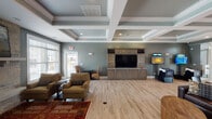This single-floor communal space is designed with both comfort and functionality in mind, offering a refined yet welcoming environment. The main living area is anchored by a striking coffered ceiling with crisp white molding, complemented by soft blue-gray walls and light hardwood-style flooring that flows throughout the space. A built-in entertainment center with custom cabinetry and a large flat-screen TV serves as the focal point of one lounge area, flanked by comfortable seating arrangements atop colorful area rugs. An adjacent seating zone features a floor-to-ceiling stone fireplace with a traditional mantle, framed by large windows that invite abundant natural light and views of the surrounding outdoor area. The kitchen is outfitted with dark wood cabinetry, granite countertops, stainless steel appliances including a French-door refrigerator and electric range, and wall-mounted TV. A casual dining nook with a round table and chairs sits beside a wide window, offering a bright and inviting spot for meals or conversation. A bar-height granite counter provides additional seating. A separate workstation with a wicker chair offers a quiet area for individual use. The overall ambiance is polished and comfortable, blending classic architectural elements with modern amenities.