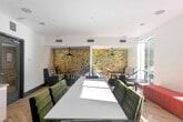 Located at 500 Race St in San Jose, California, this thoughtfully designed communal lounge space offers a compelling blend of modern aesthetics and functional versatility. The single-floor layout is unified by light-toned herringbone hardwood flooring that adds warmth and visual interest throughout.

Natural light pours in through large, floor-to-ceiling windows lining multiple walls, offering views of the surrounding streetscape and mature trees. Recessed ceiling lighting complements the abundant daylight, ensuring the space remains bright at all times.

Seating is diverse and carefully curated: plaid-upholstered sofas and gray armchairs form intimate conversation nooks, while a bold terracotta-red banquette runs along one windowed wall, paired with round café-style tables. A central white conference table with velvet green chairs provides a dedicated workspace or meeting area.

A striking multi-toned hexagonal tile feature wall in warm earth tones serves as a bold focal point, anchored by sleek pendant lights. On the opposite end, a large abstract geological artwork hangs above a built-in white cabinetry unit with a gray countertop and open shelving—ideal for storage or display.

The space is completed by a glazed entry door, ceiling-mounted speakers, and HVAC vents, reflecting a polished, amenity-rich environment suited for collaboration, socializing, or focused work.