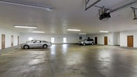This expansive enclosed garage offers a generously sized, open floor plan with a smooth concrete floor and white-painted walls that contribute to a clean, well-maintained appearance. The space is illuminated by multiple fluorescent tube lights mounted to the ceiling, ensuring bright, even coverage throughout the interior. The ceiling height is notably generous, providing ample vertical clearance for a variety of vehicles. Several small windows along the walls allow for a degree of natural light to supplement the artificial lighting. A motorized garage door opener is visible, indicating automated entry and exit access. Along the far wall, two interior doors — one painted red and one constructed of wood — suggest access to adjacent utility or storage areas. The concrete floor shows typical wear consistent with regular vehicle use. Overall, the garage presents a functional, utilitarian space well-suited for secure multi-vehicle storage, with practical amenities that enhance its everyday usability.