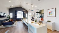 Located at 305 Cambia Dr in Schaumburg, Illinois, this contemporary single-floor community clubhouse offers a thoughtfully designed gathering space with a variety of distinct functional areas. The grand entry lobby makes an immediate impression with its soaring ceilings, stacked geometric pendant lighting, and a bold cylindrical architectural column finished in layered shades of blue and gray. Large transom windows and glass-paned entry doors flood the space with natural light.

The centerpiece of the space is an expansive open-plan lounge and kitchen area featuring vaulted ceilings, dramatic linear pendant fixtures, and an oversized white quartz island with bar seating. The adjacent kitchenette is outfitted with modern cabinetry, a stainless steel sink, and ample counter space. Dark navy built-in shelving and wainscoting accents contrast beautifully against bright white walls, lending the space a polished, upscale character.

The lounge area offers generous seating with plush sofas, accent chairs, gold-toned side tables, and a wall-mounted television — all bathed in natural light from large arched and grid-pane windows. A dedicated business center provides multiple workstations, printing capabilities, and colorful accent walls adorned with contemporary artwork, creating a productive yet vibrant workspace. Throughout, warm wood flooring and curated décor contribute to an inviting, modern ambiance.