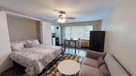 This single-floor studio at Park Chateau offers a thoughtfully designed open-concept layout that efficiently combines living, sleeping, and dining functions within one cohesive space. The main area is anchored by a built-in Murphy bed, which folds seamlessly into the wall to maximize floor space when not in use, and is complemented by a ceiling fan with integrated lighting that promotes airflow and comfort throughout. Dark hardwood flooring runs throughout the main living area, grounded by a bold geometric area rug in shades of grey, yellow, and brown that adds a lively visual focal point. A comfortable seating arrangement — including a neutral-toned sofa, accent chair, and round side table — defines the living zone, while a dedicated dining area with a table and bar-height chairs sits adjacent to a row of large windows that invite abundant natural light and offer views of surrounding greenery. The kitchen is efficiently laid out with white cabinetry, a gas range, and full-size appliances. The bathroom features classic black-and-white mosaic tile flooring, white subway wall tiles, a full soaking tub with shower, and a window that provides natural ventilation. A wall-mounted flat-screen TV and abstract artwork complete the space's functional yet inviting character.