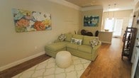This two-floor home offers a thoughtfully designed interior that balances comfort and style. The main living area immediately draws attention with its warm hardwood flooring, crisp crown molding, and neutral wall tones that create an inviting, airy atmosphere. A sectional sofa in a soft sage green anchors the living space, complemented by floral accent pillows and a round tufted ottoman atop a light geometric-patterned area rug — lending the room a fresh, relaxed character. Colorful canvas artwork adorns the walls, adding personality and visual interest to the space.

The living area flows naturally into a dining space, where a dark wood dining table and chairs provide a seamless transition between entertaining and relaxation zones. A pass-through or opening nearby hints at a connected kitchen, allowing for easy interaction between rooms. Natural light filters into the space, enhancing the warm tones of the flooring and walls.

Throughout the home, the design maintains a cohesive palette of soft neutrals accented with greens and natural wood tones, creating a harmonious and comfortable environment. Spanning two floors, the layout offers ample room for both daily living and hosting, making it a versatile and well-appointed space.