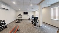 Located at 550 Park Ave W in Chatham-Kent, Ontario, this fitness center offers a well-equipped exercise space designed to support a range of workout routines. The room features a drop tile ceiling with recessed lighting that ensures bright, even illumination throughout, complemented by natural light filtering in through slatted windows along one wall. The flooring consists of durable, patterned carpet tiles suited for a high-traffic fitness environment. A wall-mounted flat-screen television is positioned prominently, providing entertainment during workouts. The cardio section is outfitted with a treadmill, a stationary spin bike, an elliptical trainer, and a recumbent exercise bike, offering varied options for cardiovascular training. A multi-station cable and weight machine provides opportunities for full-body strength training, while a dedicated free weights area includes a rack of dumbbells in multiple sizes alongside a padded workout bench. Structural columns add architectural definition to the space. A small amenity station near the entrance supplies cleaning materials and waste disposal for equipment hygiene. The room is accessed through a solid door with a window panel, with an additional door providing secondary access. Neutral-toned walls and a clean, organized layout contribute to a functional and focused exercise environment.