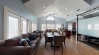 This single-floor space is designed with an open, welcoming layout that seamlessly connects its key functional areas. The centerpiece is a generous combined living and dining area anchored by soaring vaulted ceilings and a striking arched window that floods the room with natural light. A ceiling fan adds to the airy atmosphere, while rich hardwood-style flooring runs throughout, complementing the cool, calming blue-gray walls. Plantation shutters on multiple windows provide refined light control across the space.

The living area is furnished with plush, nail-head-trimmed sofas accented with teal and patterned throw pillows, creating a comfortable and inviting seating arrangement. A large dining table with upholstered chairs sits centrally, making it well-suited for gatherings. Recessed lighting and textured ceilings add further architectural character.

The kitchen is thoughtfully equipped with rich cherry-toned cabinetry, dark countertops, black appliances including a range and refrigerator, and a sink positioned beneath a shuttered window. A dedicated workstation area features a built-in counter with multiple computer setups, framed by decorative columns and benefiting from the same soaring vaulted ceiling and abundant natural light visible throughout the space.