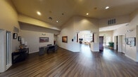 This single-story building presents a well-organized and welcoming layout designed to accommodate multiple functional areas within a cohesive open floor plan. The entry foyer greets visitors with soaring vaulted ceilings, recessed lighting, and large glass doors that flood the space with natural light, creating an immediately bright and airy atmosphere.

The main gathering area flows naturally from the entrance and features rich, dark hardwood-style flooring that extends throughout. A comfortable lounge area includes upholstered seating, a geometric accent table, and framed artwork on neutral walls, while an upright piano positioned near the entry adds a distinctive communal character.

A dedicated workstation or administrative area is situated to one side, furnished with a desk and office chair beneath recessed lighting, providing a functional workspace within the larger open plan. Arched architectural details and decorative columns add visual interest and help define transitions between areas.

The kitchenette is equipped with a full-size refrigerator, electric range, microwave, and a marble-style countertop breakfast bar with bar seating for two. A tile-surround fireplace anchors one wall of this area, adding warmth and a residential feel. Vaulted, wood-paneled ceilings in the kitchen enhance the sense of volume. A secondary seating nook, visible through an arched passage, benefits from a large arched window with views of surrounding greenery.