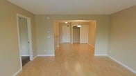 This single-floor space showcases a clean, open layout designed for comfortable everyday living. The main living area is finished with light-toned hardwood flooring that extends throughout, reflecting ambient light and contributing to a bright, airy atmosphere. Neutral beige walls provide a warm, understated backdrop that complements the flooring seamlessly.

A wide arched opening connects the living area to the entryway, creating a sense of openness and flow between spaces. The entryway is well-appointed, featuring a front door with an electronic keypad lock for added security, as well as a dedicated closet with an open rod system — ideal for coats and storage. A pendant light fixture hangs above this area, adding a decorative touch and providing focused illumination.

Additional ceiling-mounted lighting in the entryway ensures the space remains well-lit. Along the living area walls, practical features such as a thermostat panel and electrical outlets are conveniently positioned. White baseboards throughout provide a crisp contrast to the warm tones of the walls and floors, contributing to a tidy, well-finished aesthetic overall.
