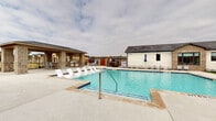 This community pool area is designed to serve as a central outdoor gathering space, centered around a generously sized swimming pool with clear, turquoise water. The pool deck is finished in light-toned concrete, providing ample space for circulation and relaxation. Numerous lounge chairs are arranged along the pool's perimeter, complemented by freestanding patio umbrellas for shade. A covered brick cabana structure on one side of the deck features an outdoor kitchen and bar area, offering a functional space for entertaining. On the opposite side, a single-story building constructed with brick and white-painted masonry houses restroom facilities, accessible via distinctive teal-painted doors. A dual-height water fountain station is conveniently located between the restroom entrances. The pool area is enclosed by a secure perimeter fence, with stone pillar accents providing architectural interest at the entry points. Surrounding views include neighboring residential homes, contributing to the community-oriented character of the space. Turf grass accents border portions of the deck, adding a landscaped touch to the overall design. The space is well-suited for both leisurely relaxation and social gatherings.