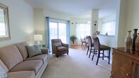 This single-floor residence offers a welcoming and functional layout designed for comfortable everyday living. The open-concept main area seamlessly connects the living room, dining space, and kitchen, creating a sense of spaciousness and ease of movement throughout.

The living room is anchored by plush wall-to-wall carpeting and furnished with a comfortable sofa and accent chair, lending a relaxed, inviting atmosphere. Soft, light green walls and neutral tones contribute to the airy feel of the space. Large sliding glass doors flood the room with abundant natural light and provide direct access to an outdoor balcony, offering pleasant views of the surrounding area.

Adjacent to the living room, a casual dining area features a bistro-style table and chairs, perfectly positioned near the sliding doors to take advantage of the natural light. A breakfast bar with barstool seating connects the dining area to the kitchen, making it an ideal spot for casual meals or conversation. The kitchen pass-through adds to the open, communicative flow between spaces.

A decorative chandelier and wall sconces add subtle elegance throughout, while the textured ceiling and clean architectural lines give the space a polished yet understated character. Overall, the residence balances practical design with a warm, comfortable ambiance.