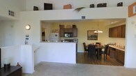 This single-floor space showcases an open-concept layout that seamlessly connects the kitchen, dining area, and living room. The kitchen features dark wood cabinetry, a black microwave, a full-size refrigerator, and a raised breakfast bar with a pass-through window that opens directly to the adjoining areas, promoting a sense of connectivity throughout the space. Upper shelving near the ceiling provides additional storage and display space, taking advantage of the notably high ceiling height. The dining area is anchored by a round dark-wood table surrounded by matching dark upholstered chairs, illuminated by an elegant chandelier that adds warmth to the space. Warm yellow-toned walls in the kitchen and dining area contrast with the neutral, light-colored walls of the living area, creating a subtle visual distinction between zones. The flooring transitions from carpet in the living area to hard flooring in the dining and kitchen spaces. A framed piece of artwork adorns the dining room wall, adding a decorative touch. The generous ceiling height throughout contributes to an airy, spacious ambiance across the entire floor plan.