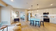 This single-floor residence offers a thoughtfully designed open-concept layout that seamlessly connects the living, dining, and kitchen areas. The kitchen is a standout feature, showcasing crisp white shaker cabinetry with under-cabinet lighting, a classic white subway tile backsplash, light quartz countertops, and a full suite of stainless steel appliances. A central island with pendant lighting provides additional prep space and casual seating for up to three, making it an ideal hub for entertaining. Light wood-style flooring flows throughout the main living areas, contributing to a bright and airy atmosphere. The living room opens to a private balcony through sliding glass doors, allowing for an abundance of natural light and outdoor access. The bedrooms are carpeted for comfort and feature generous windows with blinds that invite soft, natural light. A flexible secondary room with a sliding barn-style closet door serves well as a home office or sitting area. The bathroom is finished with white cabinetry, a clean undermount sink, and quartz countertops, maintaining the cohesive modern aesthetic found throughout the space. Overall, the residence presents a clean, neutral palette with contemporary finishes that create a welcoming and functional living environment.