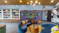 Located at 2815 Capital Blvd in Raleigh, NC, this single-story amenity space at Broadstone Oak City delivers a bold, design-forward experience defined by vibrant colors, curated artwork, and thoughtfully appointed gathering areas. The lobby commands immediate attention with a striking LED pixel-art installation displaying the community name, complemented by a warm wood-paneled ceiling, globe pendant lighting, and a curved teal reception desk. Built-in shelving with colorful accents and décor lines the walls, creating a lively yet welcoming atmosphere.

Lounge seating areas are distributed throughout, offering a mix of deep blue velvet curved sofas, retro egg chairs, and plush green settees set against exposed brick walls and patterned terrazzo flooring. Bold geometric murals and color-blocked accent walls punctuate the space, lending an artistic energy throughout.

Hallways feature distinctive black-and-white patterned encaustic tile floors framed by black-trimmed glass partitions, leading to flexible co-working or private rooms. The fitness center is well-equipped with cardio machines and strength equipment, accented by arched mirrored panels, a vibrant botanical mural, and large windows that bring in natural light and outdoor views.