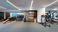 Located at 1441 Peachtree St NE in Atlanta's Midtown neighborhood, this professionally equipped fitness center offers a spacious, single-floor layout designed to accommodate a full spectrum of workout needs. The exercise room is generously proportioned, with a clean, modern aesthetic defined by white walls, recessed tray ceilings with linear LED lighting, and durable rubber flooring accented by bold blue stripes.

The cardio zone features a row of treadmills, elliptical machines, and stair climbers, all positioned near windows that allow natural light to fill the space. Large wall-mounted mirrors and flat-screen televisions enhance the environment, providing both visual depth and entertainment. Ceiling fans contribute to comfortable air circulation throughout.

The strength training area is equally well-appointed, housing multiple power racks with Olympic barbells and weight plates, a comprehensive dumbbell rack, adjustable benches, and cable machines. A functional training zone includes battle ropes, a punching bag, medicine balls, and a dedicated open floor area for dynamic exercises.

A built-in wood shelving unit with lower cabinetry and a mini-refrigerator serves as a convenience station, offering towel storage and easy access to essentials. The overall ambiance is polished and motivating, combining thoughtful design with a robust selection of equipment to support every level of fitness.