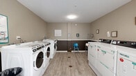 Located at 2086 Stanley Street in New Britain, Connecticut, this shared laundry facility offers a clean, efficiently organized space designed for convenience. The room is thoughtfully laid out with rows of commercial-grade front-loading washing machines along one side and a bank of top-loading dryers along the other, providing ample capacity for multiple users simultaneously. A utility sink and a wheeled laundry cart offer additional functionality for residents.

The interior features durable wood-look plank flooring in a light neutral tone, lending a clean and modern aesthetic. Walls are painted in a two-tone scheme — warm taupe on the upper half and a deep brown on the lower half, separated by a crisp white chair rail, giving the space a polished, cohesive look. A window fitted with horizontal blinds allows natural light to brighten the room, enhancing the open feel of the space.

Practical amenities include a centrally mounted flush ceiling light, a paper towel dispenser, a cork bulletin board for posted notices, and seating for those waiting. The room is well-maintained and thoughtfully appointed, reflecting a commitment to providing residents with a comfortable and fully functional shared laundry experience.