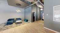 Located on Prospect Avenue in Cleveland, Ohio, Innovation Landing is a thoughtfully designed single-floor collaborative space that blends industrial aesthetics with modern comfort. The open-concept layout features exposed ductwork and structural columns alongside a striking sculptural ceiling installation — a dramatic wave of layered navy blue fins that serves as the visual centerpiece of the space.

The flooring transitions between light-toned hexagonal wood-look tile and patterned carpet tiles, subtly delineating different functional zones throughout. A central high-top table with metal stools anchors a casual gathering area, while multiple lounge configurations — including a warm caramel sofa, teal armchairs, and vibrant yellow accent seating — offer flexible options for collaboration or relaxation.

Floor-to-ceiling windows along the exterior wall flood the space with natural light and provide open views of the surrounding Cleveland streetscape. A long wooden bar-height counter with slender chairs sits adjacent to the window wall beneath elegant pendant lighting, ideal for focused work or informal meetings. A pool table adds a recreational element to the far end of the room. Dark-framed doors with glass panels lead to adjoining areas, and a cozy lounge nook with a sofa and pendant cluster light fixture provides a quieter retreat within the space.