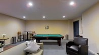This single-floor recreational space is designed as a casual lounge and games room. The centerpiece is a full-sized pool table with a green felt surface and dark wood frame, positioned prominently within the room. Recessed ceiling lights provide even, ambient illumination throughout the space, creating a well-lit environment suitable for gameplay and socializing.

The seating area includes a light-toned sofa adorned with decorative pillows and a dark leather armchair, arranged to encourage conversation and relaxation. A tall bistro-style table with a chair is positioned along one wall, offering an additional spot for casual seating or spectating.

The walls are finished in a warm, neutral tone, contributing to the room's inviting ambiance. The flooring features a combination of carpet and hard surface areas, adding visual texture. A small piece of framed artwork on the wall adds a subtle decorative touch. A metal waste bin is neatly placed near the pool table, reflecting a functional and tidy layout. Overall, this space is well-suited for informal gatherings and recreational use.