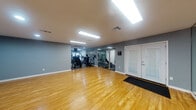 This single-floor fitness center is thoughtfully designed to accommodate a wide range of workout styles and fitness goals. The main open area features polished hardwood flooring that provides ample space for stretching, yoga, aerobics, or group exercise sessions. Full-length mirrors are mounted along the walls, enhancing the sense of space while allowing users to monitor their form during workouts. The space is illuminated by recessed lighting and flush-mount fluorescent fixtures, ensuring bright, even lighting throughout.

The strength training zone transitions to durable rubberized flooring and is anchored by a commercial-grade cable machine with multiple attachment points, ideal for a comprehensive full-body workout. A well-stocked dumbbell rack lines the mirrored wall, offering a range of free weight options. A dedicated cardio area houses multiple machines, including an elliptical trainer, a stationary bike, and a treadmill, catering to varied cardiovascular training preferences.

Additional equipment includes a medicine ball rack and stability balls, supporting functional fitness and core training. The muted gray walls create a focused, professional atmosphere, while a glass-paneled entry door adds a polished touch. A small utility area with a sink is also present, adding convenience for users. Overall, this fitness center delivers a complete and functional exercise environment.