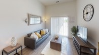 This single-floor unit showcases a contemporary design with notably high ceilings that lend an airy, open feel throughout. The living area features wood-style plank flooring and is anchored by a large sliding glass door fitted with vertical blinds, allowing generous natural light and outdoor views. The space is well-proportioned, comfortably accommodating a full seating arrangement and entertainment setup.

The kitchen is efficiently laid out and well-appointed, featuring dark espresso shaker-style cabinetry paired with granite countertops and a classic white subway tile backsplash. A full suite of stainless steel appliances includes a refrigerator, electric range, built-in microwave, and dishwasher, providing a highly functional cooking environment.

Two bedrooms offer comfortable, carpeted retreats, each with natural light from windows dressed in sheer curtains. One bedroom includes a wall-mounted television, while both rooms provide a calm, neutral atmosphere suited for rest.

A dedicated dressing or storage corridor connects the bedroom area to the bathroom, which mirrors the kitchen's design language with dark cabinetry, granite countertops, a large vanity mirror, and a tub-shower combination. Light wood-style flooring and brushed nickel fixtures complete the polished, cohesive aesthetic throughout the unit.