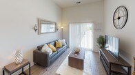 This single-floor unit showcases a contemporary design with notably high ceilings that lend an airy, open feel throughout. The living area features wood-style plank flooring and is anchored by a large sliding glass door fitted with vertical blinds, allowing generous natural light and outdoor views. The space is well-proportioned, comfortably accommodating a full seating arrangement and entertainment setup.

The kitchen is efficiently laid out and well-appointed, featuring dark espresso shaker-style cabinetry paired with granite countertops and a classic white subway tile backsplash. A full suite of stainless steel appliances includes a refrigerator, electric range, built-in microwave, and dishwasher, providing a highly functional cooking environment.

Two bedrooms offer comfortable, carpeted retreats, each with natural light from windows dressed in sheer curtains. One bedroom includes a wall-mounted television, while both rooms provide a calm, neutral atmosphere suited for rest.

A dedicated dressing or storage corridor connects the bedroom area to the bathroom, which mirrors the kitchen's design language with dark cabinetry, granite countertops, a large vanity mirror, and a tub-shower combination. Light wood-style flooring and brushed nickel fixtures complete the polished, cohesive aesthetic throughout the unit.