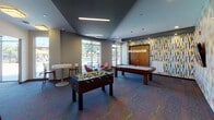 Located at 6920 Meadowbrook Blvd in St. Louis Park, Minnesota, this single-floor amenity space is designed as a lively and engaging recreation lounge. The game room serves as the centerpiece, anchored by a full-size pool table beneath a stylish pendant light fixture and a foosball table, both set atop a bold, multicolored patterned carpet that adds energy to the space. A vibrant, multi-hued geometric accent wall provides a striking visual backdrop, complemented by a wall-mounted flat-screen television. Comfortable red swivel chairs and high-top bistro tables offer casual seating throughout. An arcade game station is situated near the entryway, adding a nostalgic touch. Large floor-to-ceiling windows and glass doors flood the space with natural light while framing views of an outdoor pool area complete with lounge seating and umbrellas, creating a seamless indoor-outdoor connection. The ceiling features a combination of recessed lighting and linear flush-mount fixtures set against a dark charcoal finish, lending a modern, polished aesthetic. A dedicated area adjacent to the game room appears to function as a lounge or reading nook, with shelving visible through a glass partition. The overall ambiance is contemporary, colorful, and social — purpose-built for community recreation and relaxation.