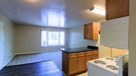 This single-floor space showcases an open-concept design that seamlessly connects the kitchen and living area. The kitchen is equipped with warm medium-toned wood cabinetry, both upper and lower, paired with dark countertops that provide a pleasing contrast. A peninsula-style counter extends outward, creating a natural divider between the kitchen and the adjoining living space while offering additional prep and storage space. A white electric range and refrigerator complete the kitchen's functional layout, and a flush-mount fluorescent light fixture provides direct overhead illumination.

The transition from the kitchen's hard flooring to the carpeted living area adds textural variety throughout the space. The living area benefits from a wide window that draws in natural light and offers a view of the surrounding greenery outdoors, contributing to an airy and open atmosphere. Neutral-toned walls complement the overall palette, keeping the interior feeling clean and versatile. The layout is practical and straightforward, making efficient use of the available floor space while maintaining a comfortable flow between functional zones.