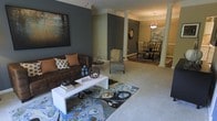 This well-appointed single-floor residence offers an open and flowing layout that seamlessly connects the living and dining areas. The living room is anchored by a rich brown leather sofa accented with warm earth-toned throw pillows, complemented by a sleek white coffee table and a vibrant blue and earth-toned area rug that adds a striking focal point to the space. Deep slate-gray accent walls provide a bold, modern contrast against lighter neutral tones, while large decorative artwork enhances the sophisticated ambiance. An elegant structural column frames the transition between the living and dining areas, adding a classic architectural detail to the open-concept design. The dining area features a dark round table with matching chairs beneath a statement pendant light, creating an intimate yet refined setting. Crown molding along the ceilings reinforces the upscale character of the space. Plush carpeting throughout adds comfort underfoot, while the warm, layered lighting creates an inviting atmosphere across all areas. The overall design balances contemporary style with classic architectural elements, resulting in a cohesive and thoughtfully curated living environment.