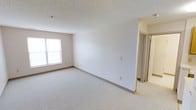 This single-floor space presents a straightforward, functional layout with a clean and neutral aesthetic throughout. The main living area is finished with light-toned carpet flooring that extends across the room, providing a soft underfoot feel. Walls are painted in a crisp white, lending a bright and open atmosphere to the interior. A textured popcorn ceiling runs overhead, a characteristic finish commonly found in residential construction.

Natural light enters the space through a double-hung window positioned along one wall, offering a source of ambient daylight that enhances the room's openness. Standard electrical outlets and switches are distributed at practical intervals along the walls, reflecting a utilitarian design approach.

A hallway extends from the main room, providing access to additional areas within the unit. The corridor features the same neutral wall tones and transitions toward what appears to be a tiled space, likely a bathroom or utility area, partially visible through an open door.

Overall, the space offers a blank canvas with minimal decorative elements, making it well-suited for customization. Its clean finishes, practical layout, and neutral palette provide a versatile foundation for a variety of uses and furnishing styles.