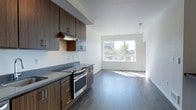 This single-floor space showcases a thoughtfully designed open-concept layout where the kitchen flows seamlessly into the living area. The kitchen is outfitted with rich, dark-toned wood cabinetry featuring sleek metal hardware, complemented by grey stone countertops that provide ample workspace. A stainless steel range with a built-in overhead ventilation hood serves as a functional centerpiece, while additional upper and lower cabinetry offers generous storage. The countertop extends to accommodate a cooktop, maximizing utility within the space. Consistent grey-toned hardwood flooring runs throughout, visually connecting the kitchen and living areas while adding warmth and continuity to the overall design. The living area is bright and open, benefiting from a large picture window that invites abundant natural light and frames views of the surrounding outdoor environment. Clean white walls and a crisp white ceiling enhance the sense of openness throughout. The overall aesthetic is modern and minimalist, combining functional design elements with contemporary finishes to create a cohesive, light-filled living environment.