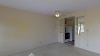 This single-floor space offers a straightforward, functional layout designed for comfortable everyday living. The main living area is finished with wall-to-wall carpeting in a neutral tone, contributing to a clean and understated aesthetic. A ceiling fan with an integrated light fixture is centrally mounted overhead, providing both ventilation and ambient lighting throughout the room. The walls are painted in a soft, warm beige, lending the space a modest, inviting feel. Natural light enters through what appears to be a glass-block window, adding a subtle architectural detail while maintaining privacy.

Adjacent to the living area, the kitchen is accessible through an open doorway and is equipped with essential appliances, including a refrigerator, a range with oven, a microwave, and additional cabinetry for storage. The kitchen features darker flooring that contrasts with the carpeted living space, helping to visually define the two areas. A wall-mounted ventilation unit is visible in the kitchen, ensuring adequate air circulation. Overall, the space presents a practical, no-frills environment suitable for single or small-household occupancy.