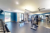 Located at 9300 Lottsfull Rd in Upper Marlboro, MD, this fitness center offers a well-appointed, single-floor layout across a generously sized footprint, divided into two distinct exercise rooms designed to support a wide range of workout needs.

The primary exercise room is bright and open, featuring speckled rubber flooring, ceiling fans, and large windows that invite natural light and outdoor views. This space is equipped with a row of treadmills, elliptical machines, and a variety of strength-training stations, including cable machines, a power rack with bench press, and free weights. Bold blue accent walls and a motivational fitness-themed wall graphic add an energetic atmosphere throughout. A water station with a hydration dispenser is conveniently positioned along the wall. An interior staircase with modern stainless steel railings connects sections of the facility.

The second exercise room serves as a dedicated studio space, featuring warm hardwood flooring and a clean, minimalist design with neutral gray walls. Floor-to-ceiling windows and glass doors provide direct access to the outdoors, filling the room with natural light. This versatile studio accommodates cycling bikes, balance trainers, and a wall-mounted television, making it well-suited for group fitness classes or individual training. Together, the two rooms create a comprehensive and functional fitness environment.