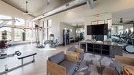 Located at 1035 N Day Miar Rd in Grand Prairie, Texas, this single-floor fitness center is designed to accommodate a wide range of workout styles and fitness goals. The space is defined by soaring ceilings and expansive floor-to-ceiling windows framed in black, which flood the interior with natural light and offer views of the surrounding exterior stonework and outdoor environment.

The facility is organized into distinct workout zones. A cardio area near the entrance houses a row of modern treadmills positioned alongside a lounge space with wood-framed seating and patterned carpet tiles, creating a welcoming transition between relaxation and exercise. Exposed ductwork runs overhead, lending an industrial aesthetic to the high-ceilinged interior.

A dedicated strength and functional training zone features cable machines, weight benches, a full dumbbell rack with mirrored walls, and a large mounted television. A separate section accommodates specialty equipment including a rowing machine, medicine ball rack, and a heavy punching bag, set against dark hardwood-style flooring. The entrance features black double glass doors flanked by limestone-clad walls, reinforcing the modern yet warm architectural character throughout.