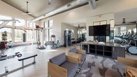 Located at 1035 N Day Miar Rd in Grand Prairie, Texas, this single-floor fitness center is designed to accommodate a wide range of workout styles and fitness goals. The space is defined by soaring ceilings and expansive floor-to-ceiling windows framed in black, which flood the interior with natural light and offer views of the surrounding exterior stonework and outdoor environment.

The facility is organized into distinct workout zones. A cardio area near the entrance houses a row of modern treadmills positioned alongside a lounge space with wood-framed seating and patterned carpet tiles, creating a welcoming transition between relaxation and exercise. Exposed ductwork runs overhead, lending an industrial aesthetic to the high-ceilinged interior.

A dedicated strength and functional training zone features cable machines, weight benches, a full dumbbell rack with mirrored walls, and a large mounted television. A separate section accommodates specialty equipment including a rowing machine, medicine ball rack, and a heavy punching bag, set against dark hardwood-style flooring. The entrance features black double glass doors flanked by limestone-clad walls, reinforcing the modern yet warm architectural character throughout.